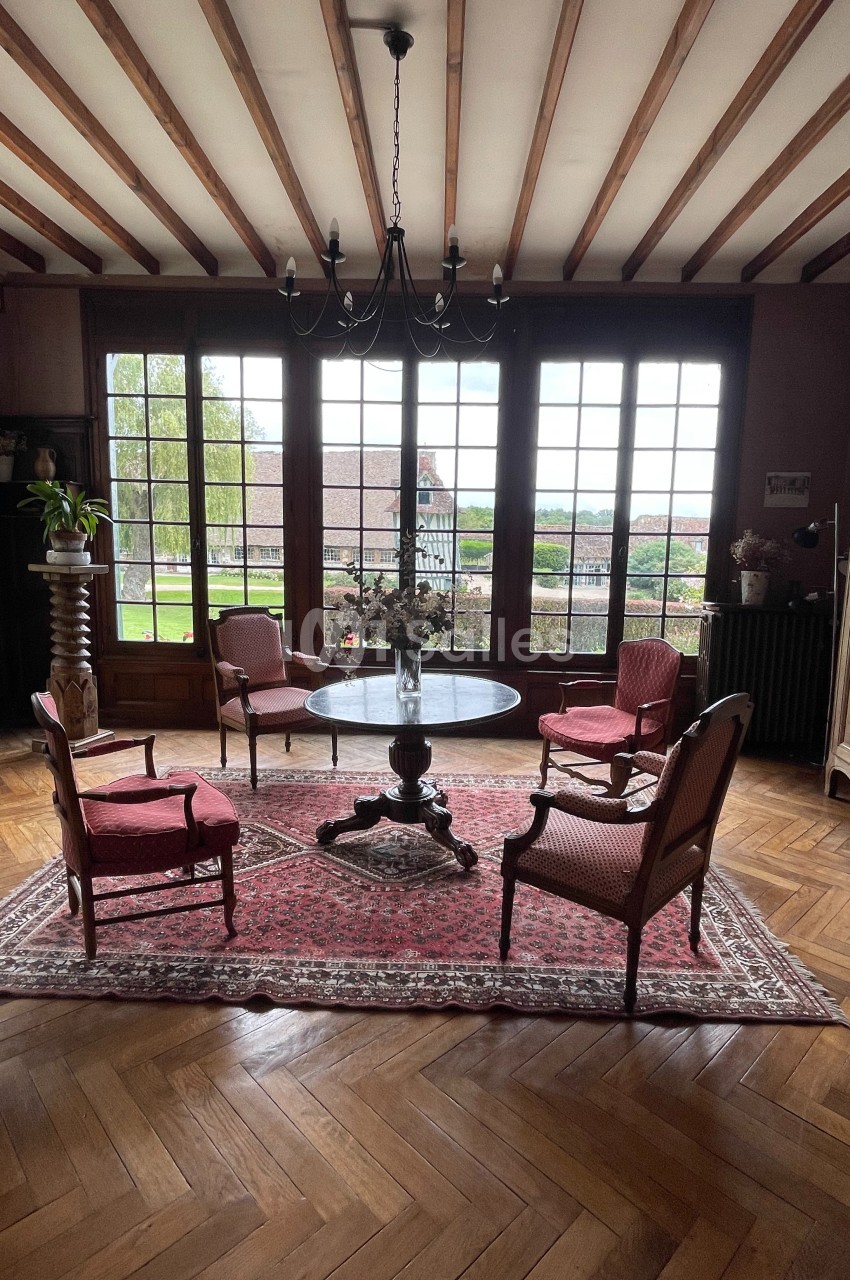 Salon avec parquet en bois, table ronde, quatre fauteuils rouges, tapis, chandelier et grandes fenêtres donnant sur un…
