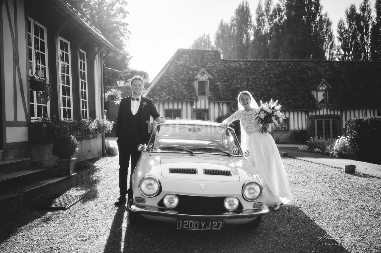 Un couple en tenue de mariage pose près d'une voiture ancienne devant une maison à colombages.