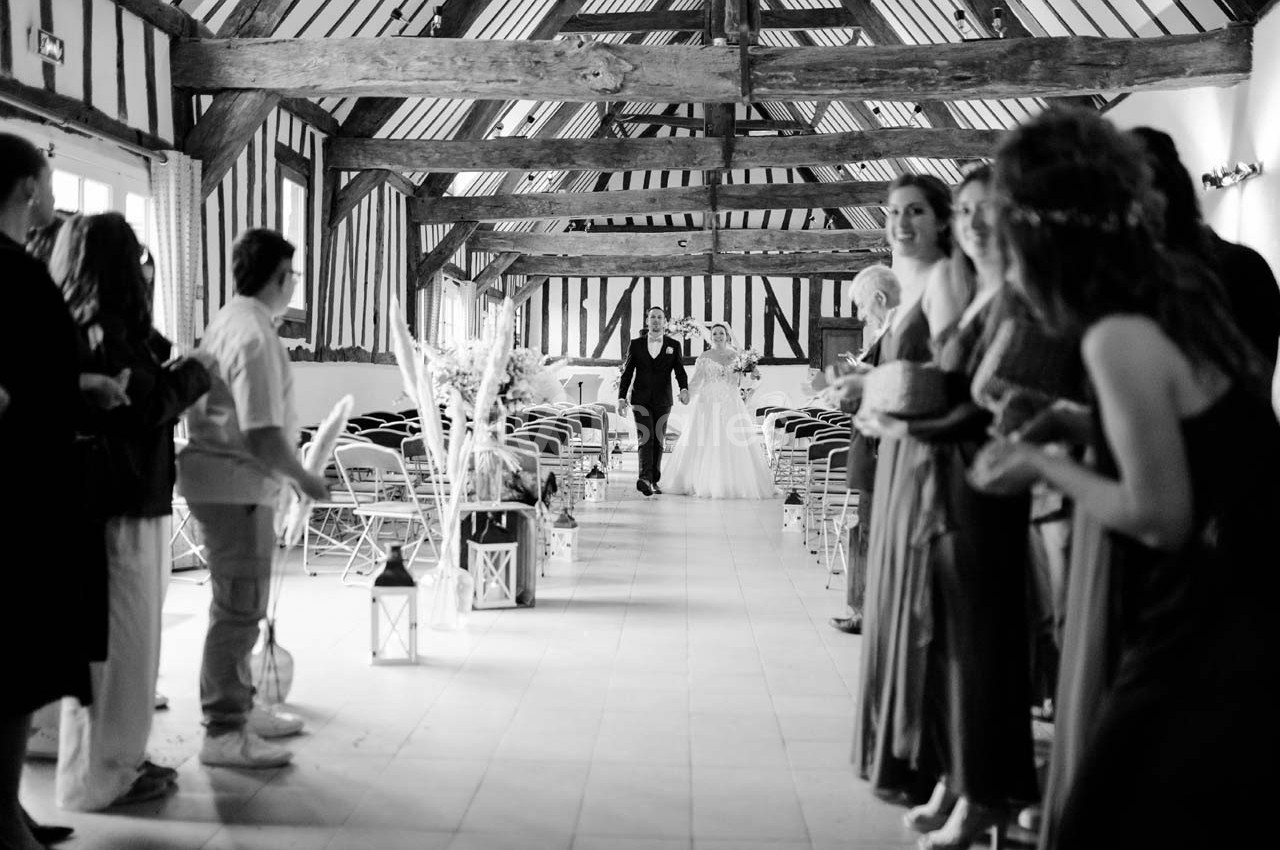 Un couple avance dans une allée centrale d'une salle en bois, entouré d'invités debout applaudissant.