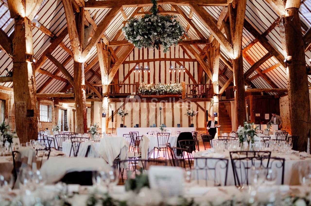Salle de réception rustique avec poutres apparentes, tables décorées et un grand lustre floral suspendu.
