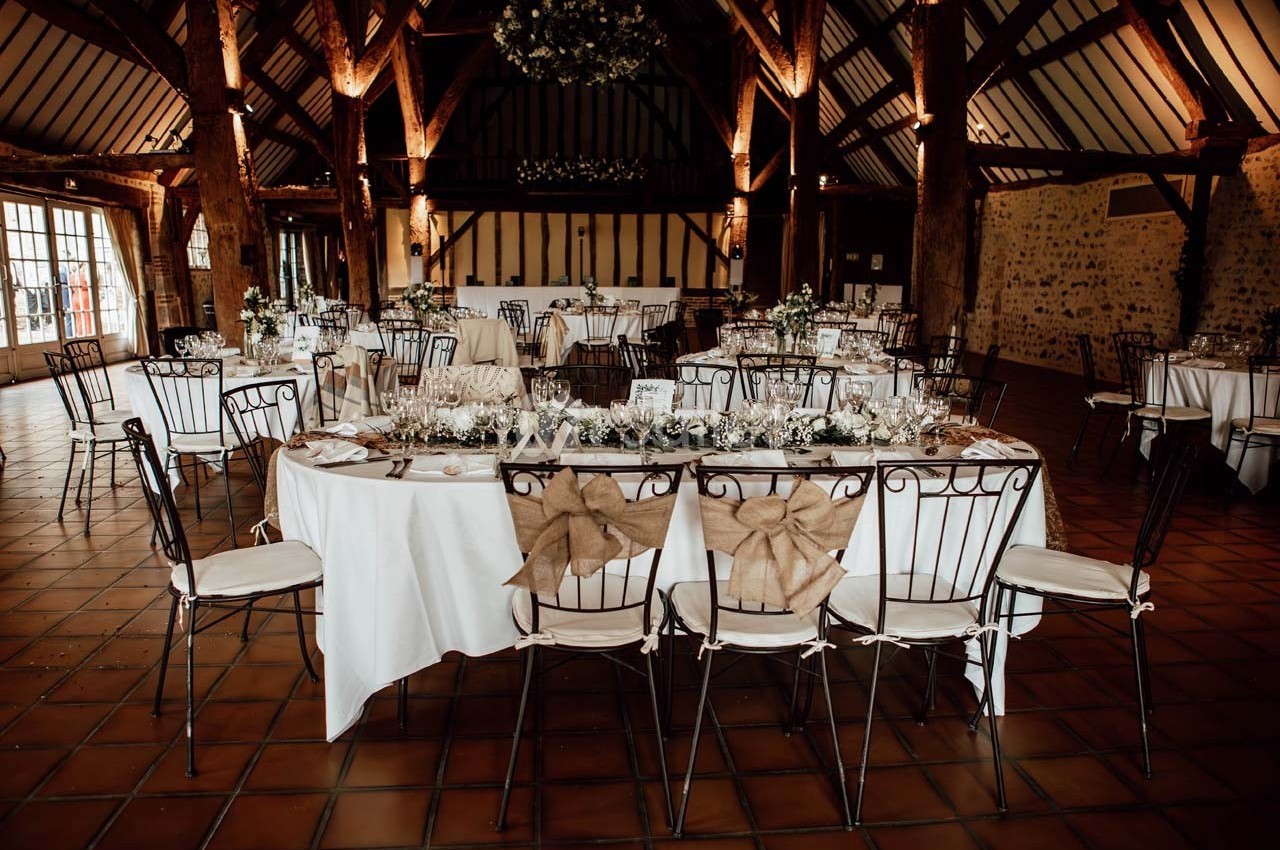 Salle de réception avec tables décorées, chaises ornées de nœuds en toile de jute et poutres apparentes en bois.