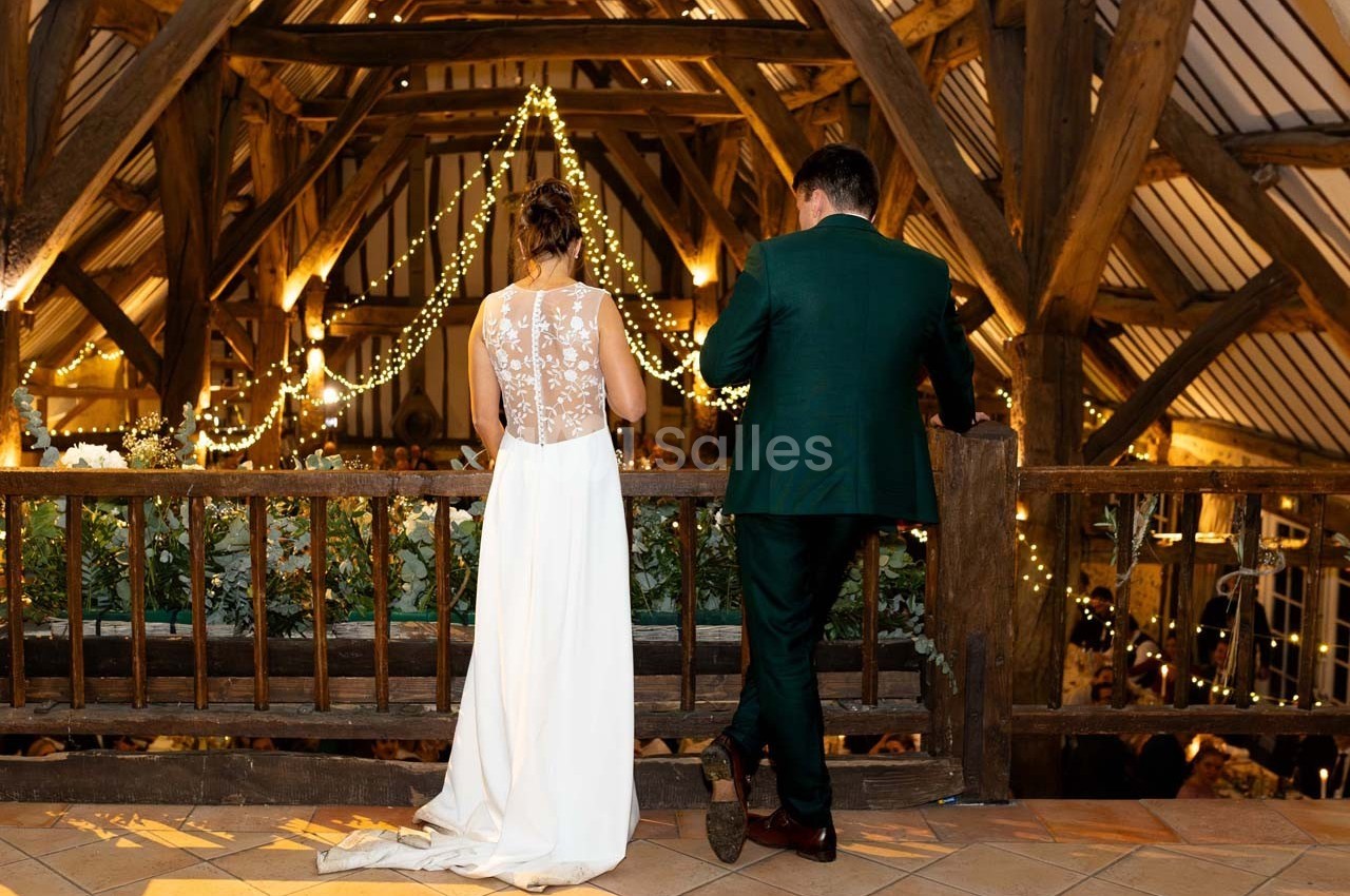 Un couple en tenue de mariage se tient sur une mezzanine en bois décorée de guirlandes lumineuses.