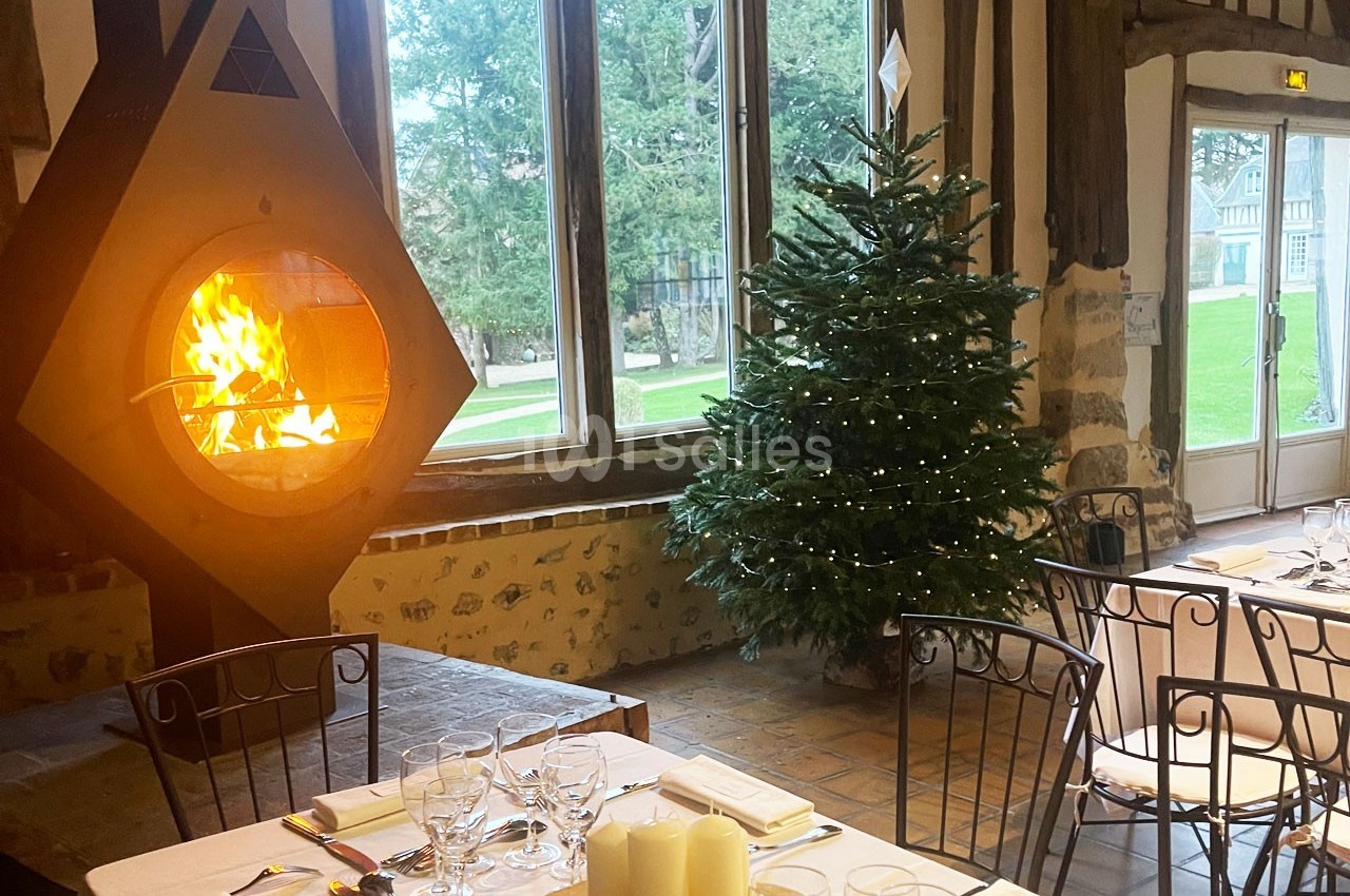 Salle de restaurant chaleureuse avec cheminée allumée, sapin décoré et tables dressées près de grandes fenêtres.