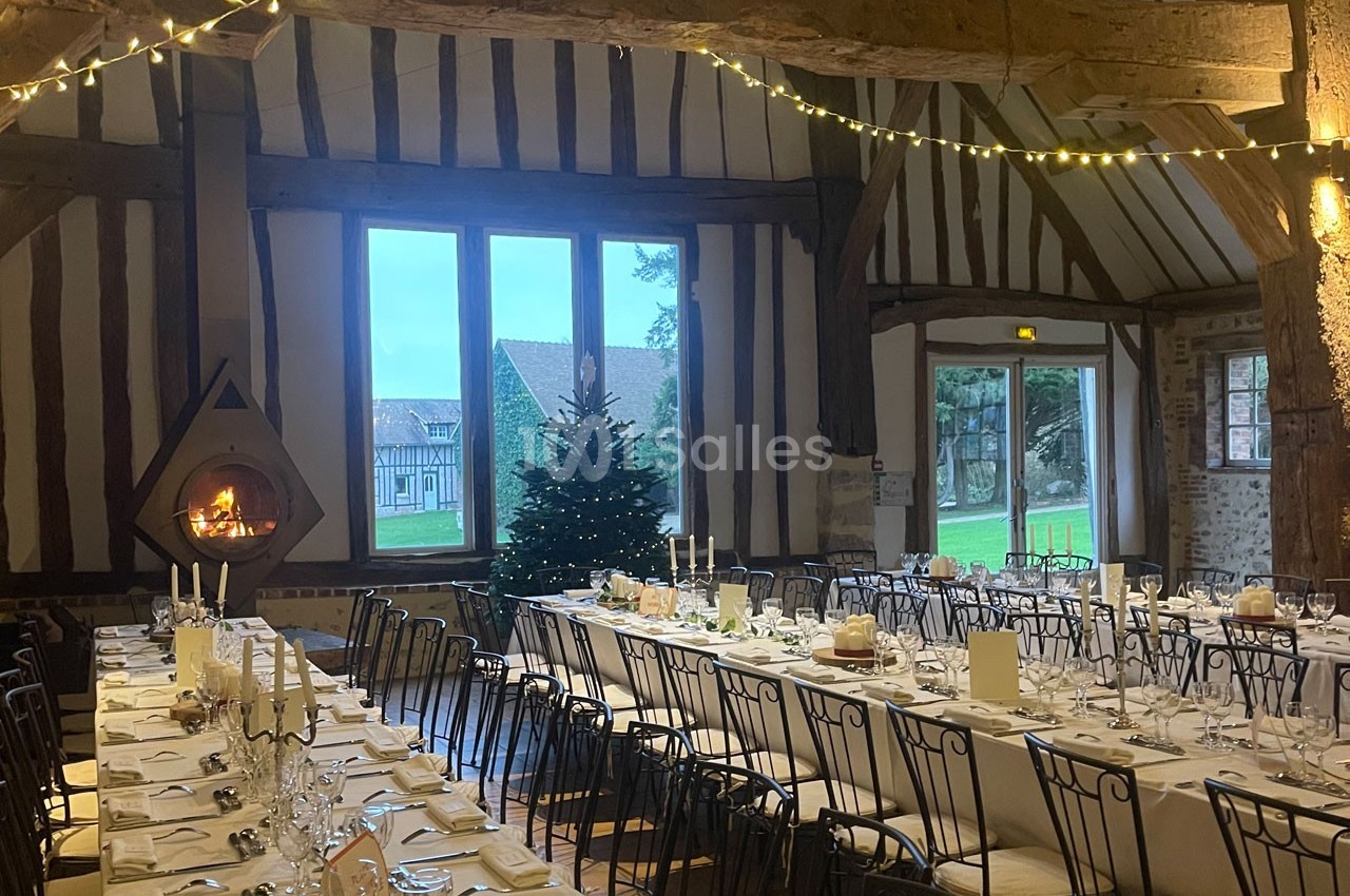 Salle de réception rustique avec poutres apparentes, tables dressées, cheminée allumée et sapin décoré près d'une fenêtre.