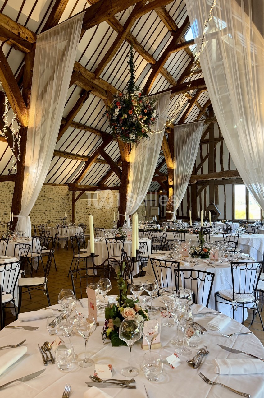 Salle de réception rustique avec poutres apparentes, tables rondes dressées et décoration florale élégante.