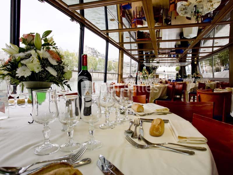 Salle de restaurant sur une péniche avec tables élégamment dressées, fleurs et vue sur la Seine à travers des baies vitrées.