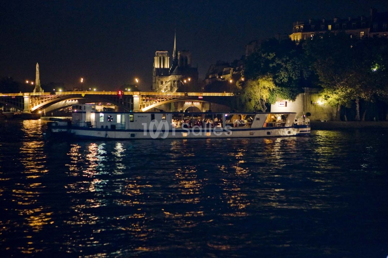 Bateau illuminé naviguant de nuit sur la Seine, avec Notre-Dame et un pont éclairé en arrière-plan.