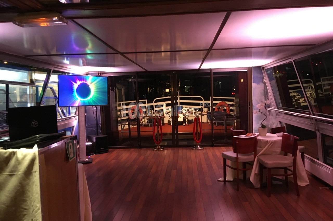 Salle intérieure d'un bateau avec parquet en bois, écran lumineux, table dressée et vue sur le pont extérieur.