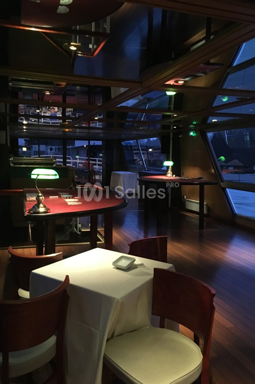 Salle intérieure d'un bateau avec des tables en bois, nappes blanches, éclairage tamisé et vue sur l'extérieur.