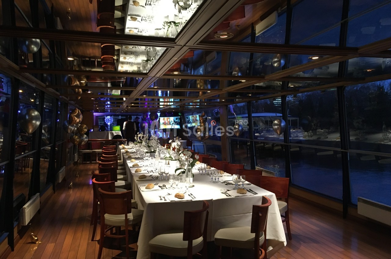 Salle de réception élégante avec grandes baies vitrées, tables dressées et vue nocturne sur l'extérieur.