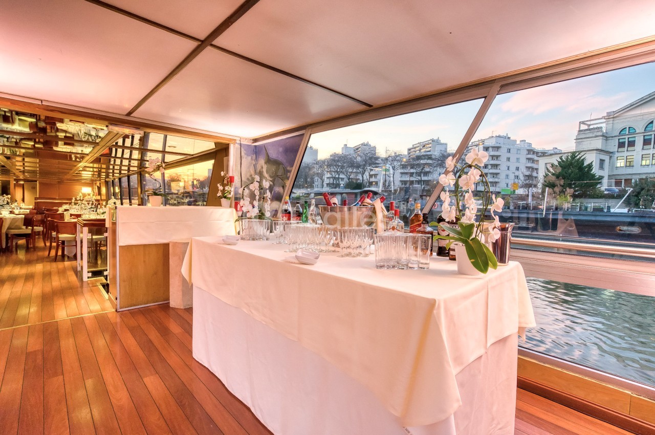 Salle de réception sur un bateau avec vue sur l'eau, table dressée avec verres, seaux à champagne et décorations florales.