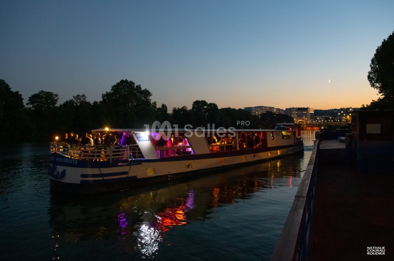 Bateau éclairé naviguant sur une rivière au crépuscule, avec des passagers visibles à bord.