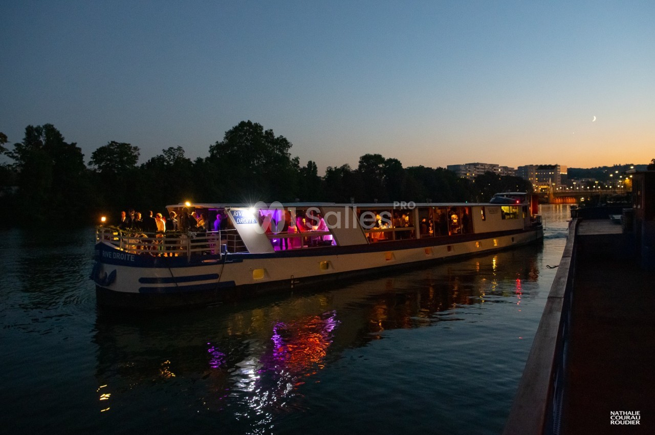 Bateau illuminé naviguant sur une rivière au crépuscule, avec des passagers visibles sur le pont.