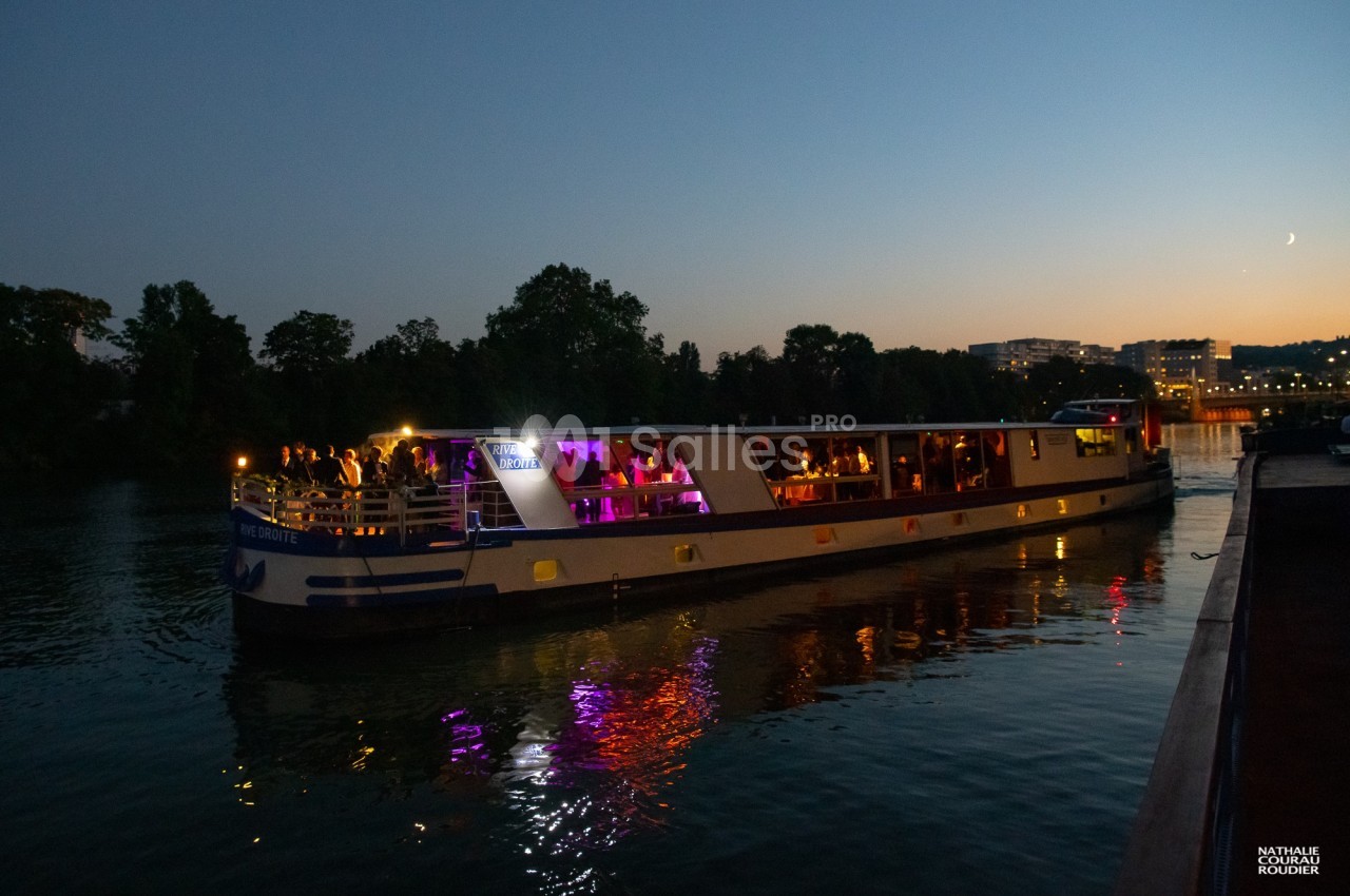 Bateau illuminé naviguant sur une rivière au crépuscule, avec des passagers visibles à bord.