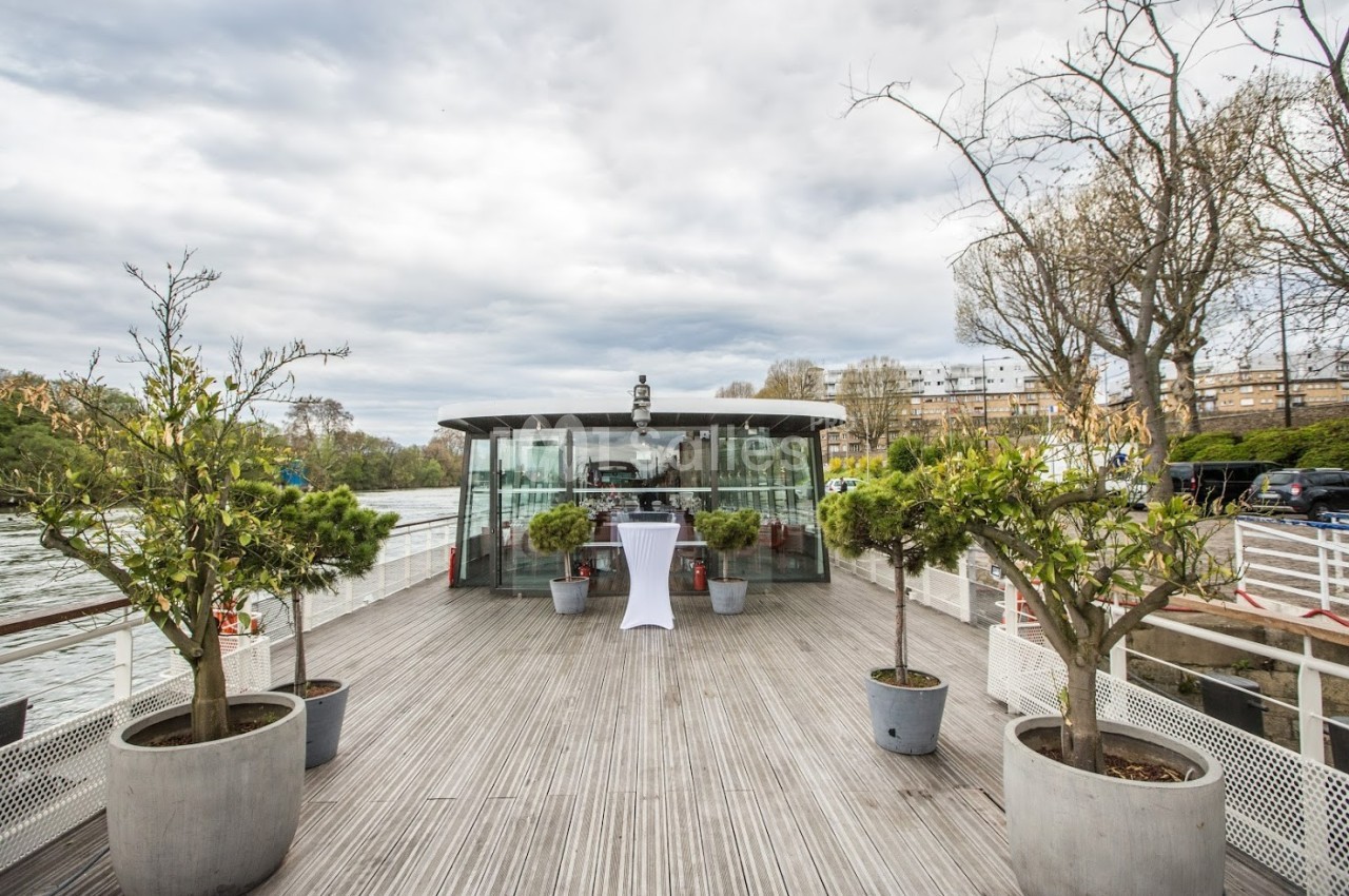 Terrasse en bois d'un bateau avec des plantes en pot, vue sur une salle vitrée et un fleuve en arrière-plan.