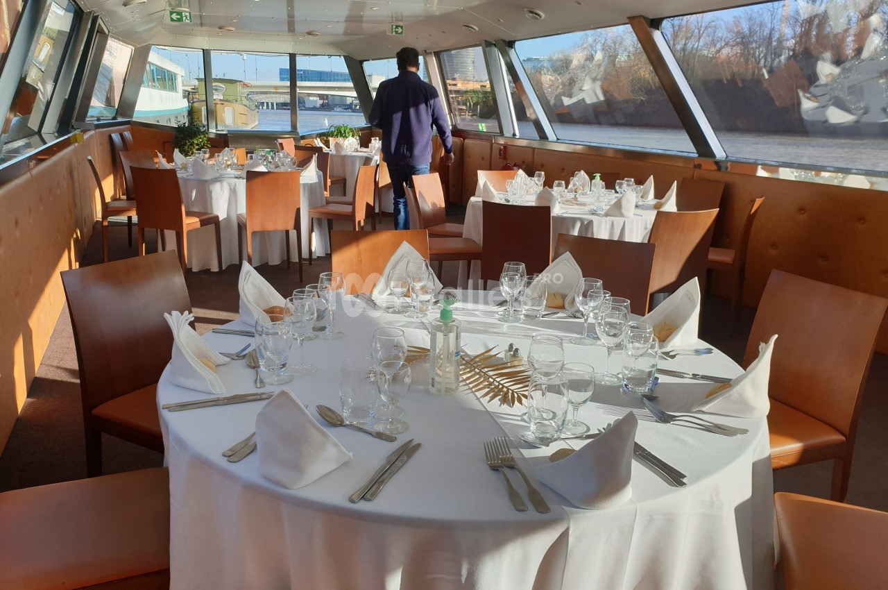 Salle de restaurant sur un bateau avec tables rondes dressées, nappes blanches et vue sur un paysage fluvial.