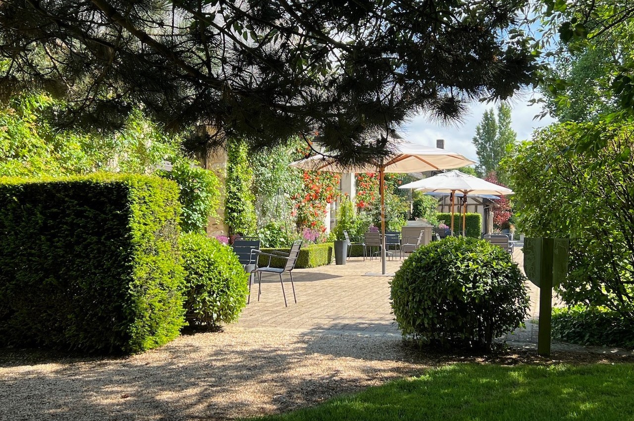 Jardin avec terrasse ombragée, mobilier de jardin, parasols et haies taillées sous un ciel partiellement ensoleillé.