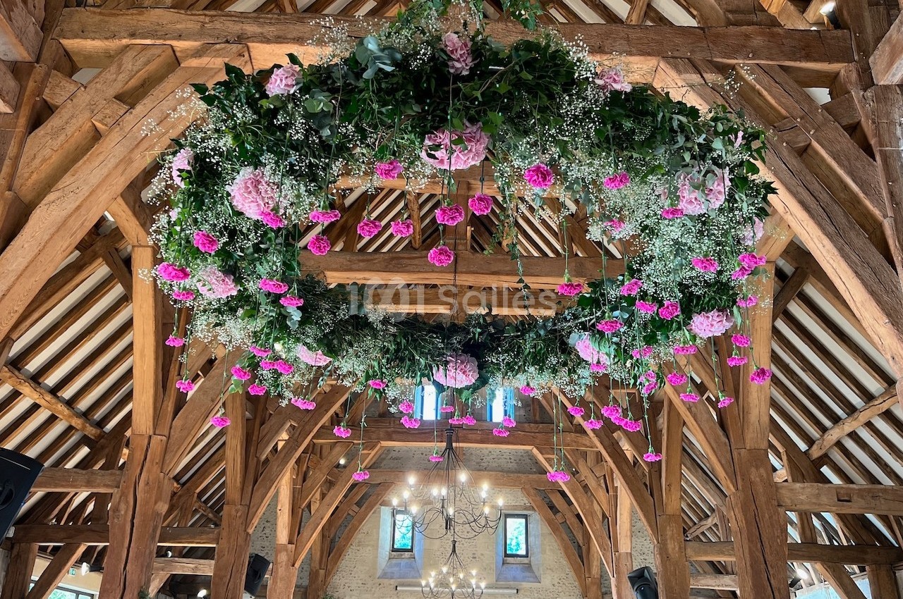 Couronne suspendue de fleurs roses et feuillage, décorant une charpente en bois dans une salle lumineuse.