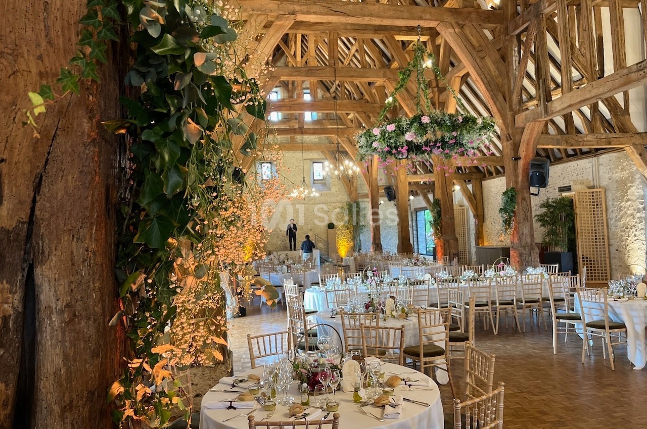 Salle de réception rustique avec poutres apparentes, tables décorées et guirlandes florales suspendues.