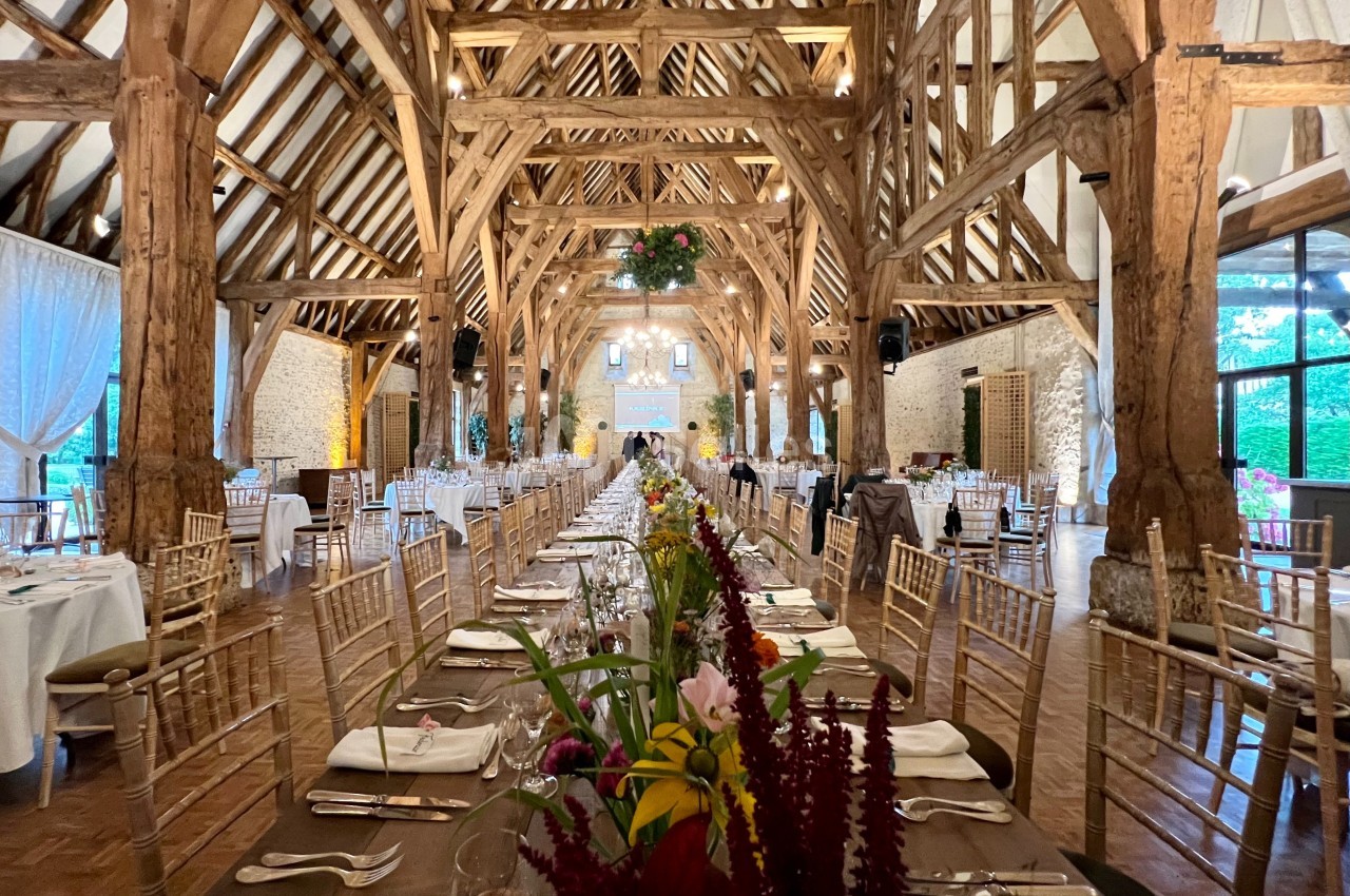 Grande salle de réception avec charpente en bois apparente, tables dressées et décor floral au premier plan.