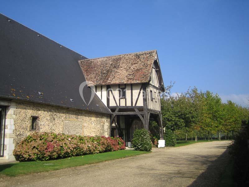 Bâtiment à colombages avec toit en tuiles, entouré de haies fleuries et d'une allée gravillonnée sous un ciel dégagé.