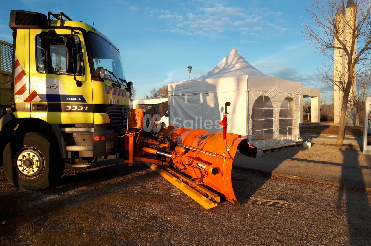 Camion équipé d'une lame de déneigement orange stationné près d'une tente blanche sur un terrain dégagé.