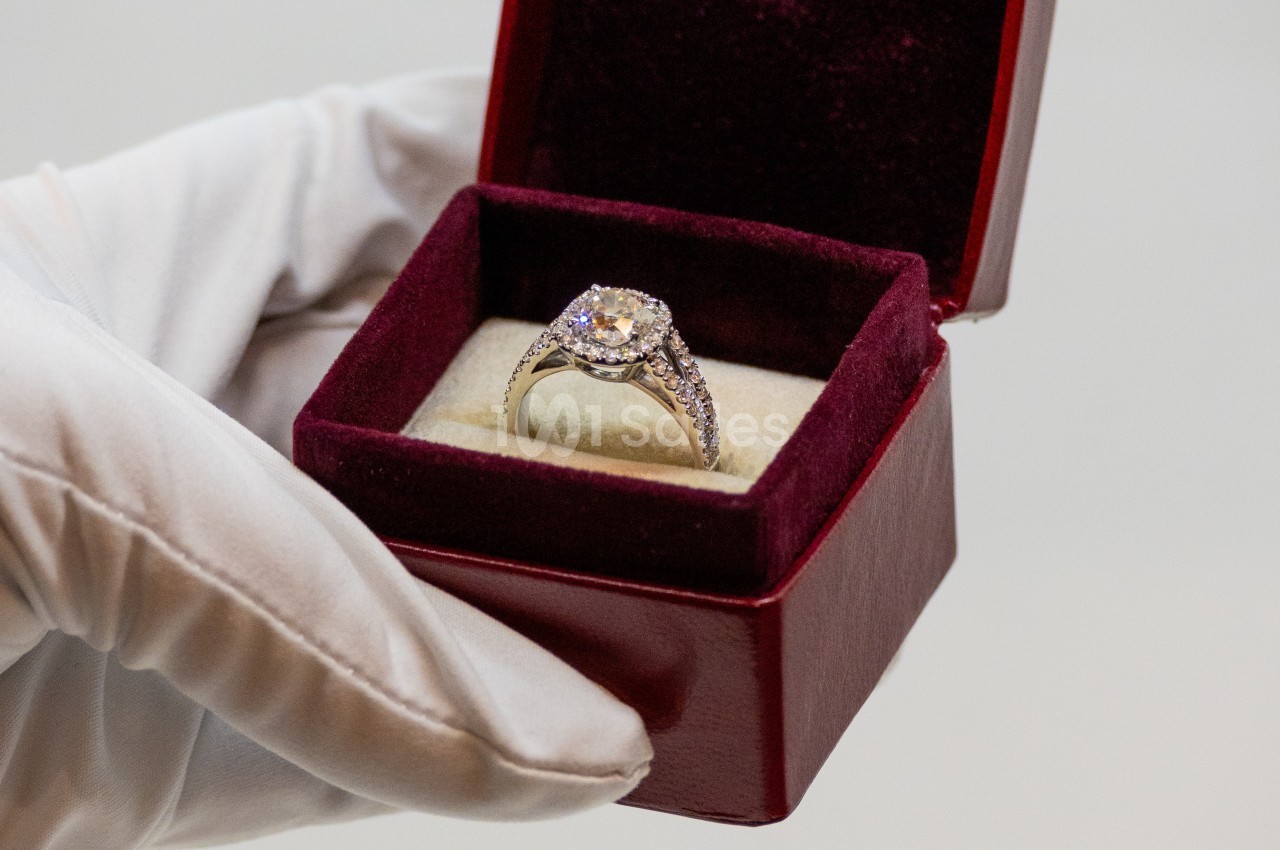 Bague ornée d'un diamant central et de petits diamants, présentée dans un écrin rouge par une main gantée.