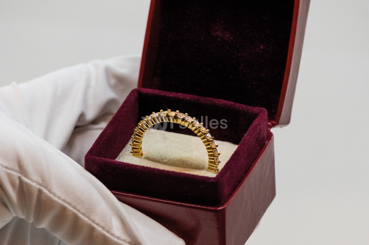 Une main gantée tient un écrin ouvert contenant une bague en or ornée de petites pierres.