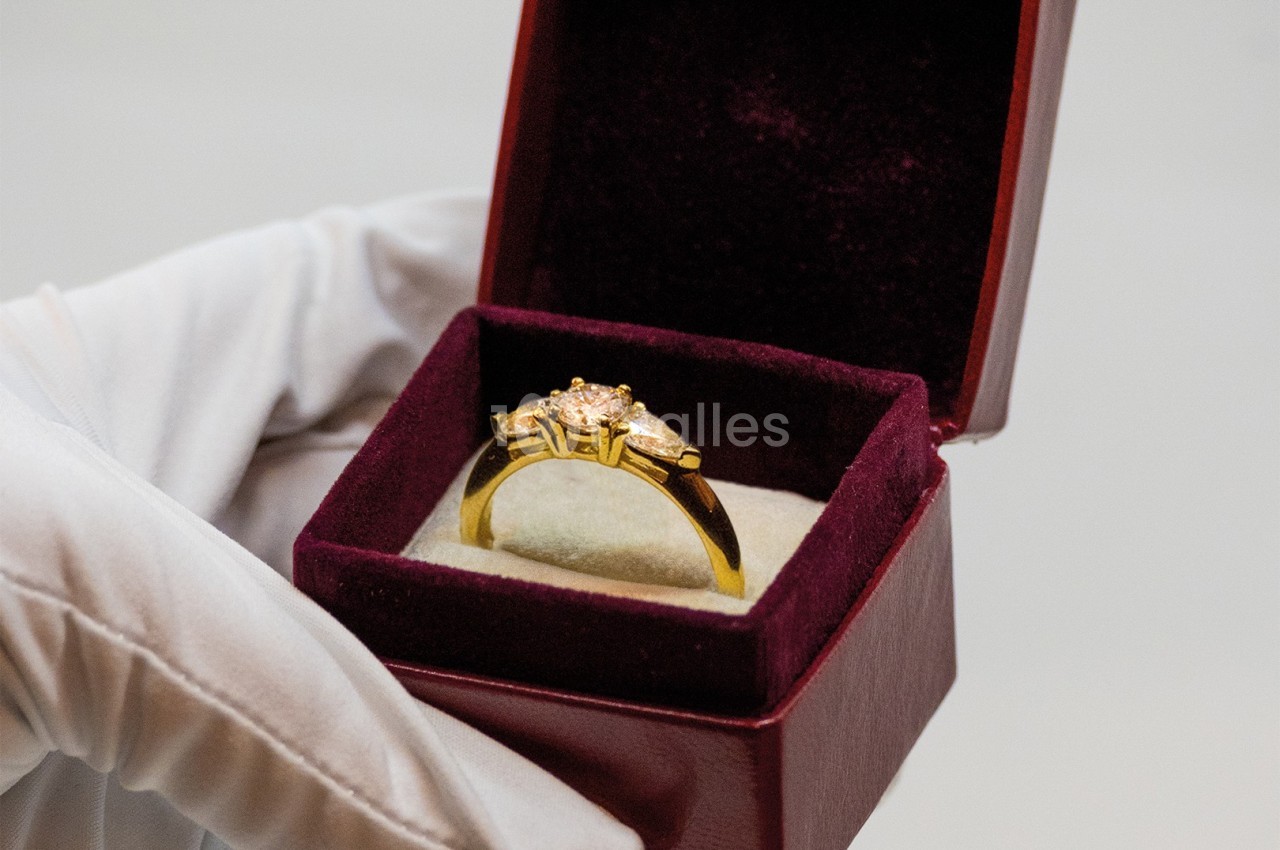 Bague en or ornée de pierres précieuses dans un écrin rouge, tenue par une main gantée blanche.