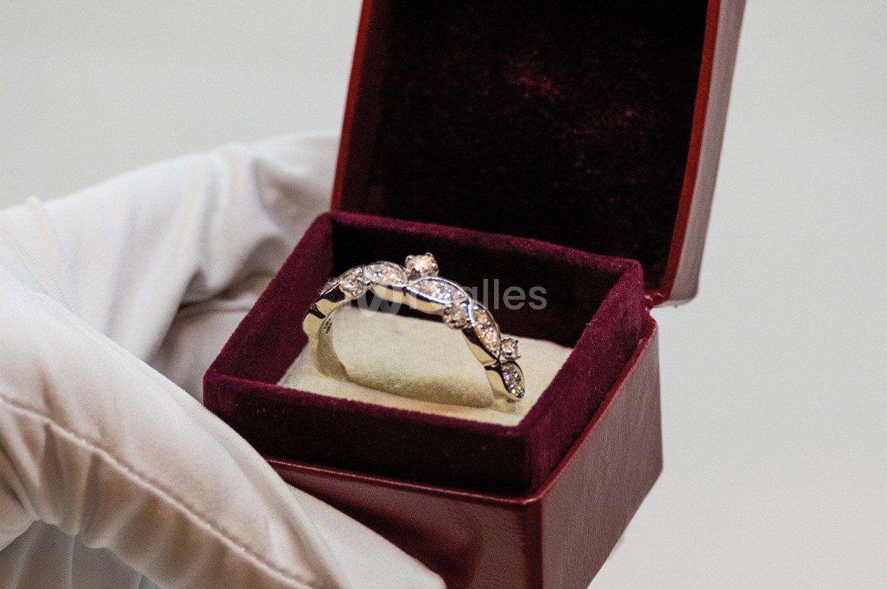 Une main gantée tient une boîte ouverte contenant une bague ornée de pierres précieuses.