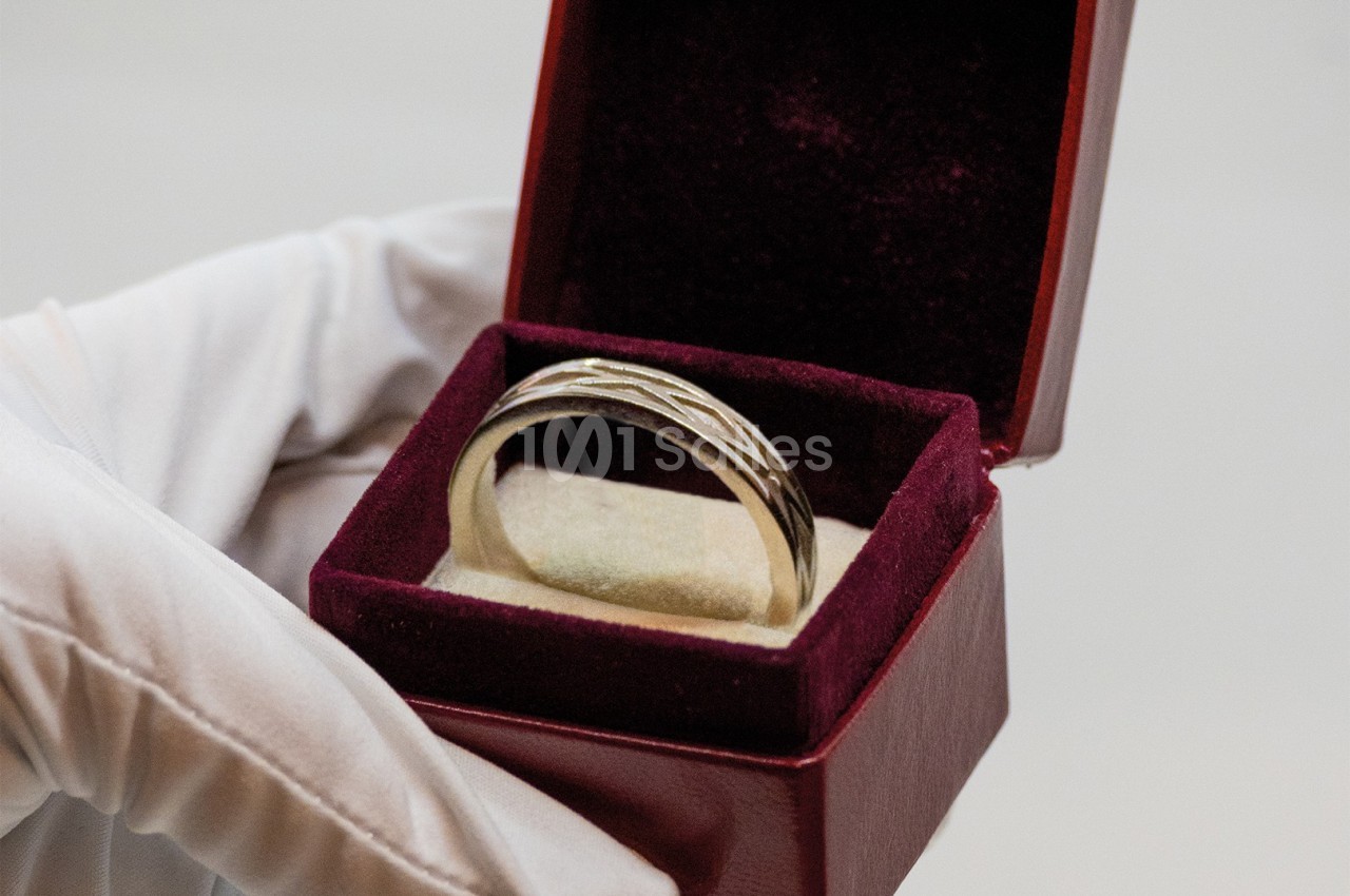 Une main gantée tient un écrin ouvert contenant une bague argentée sur un coussin en velours.