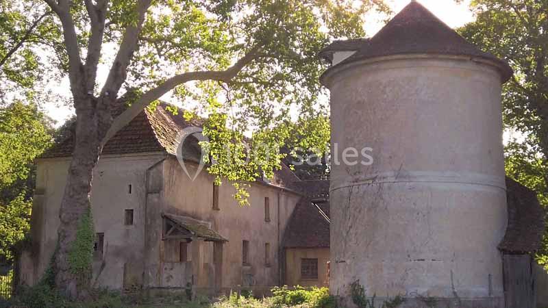 Bâtiment ancien en pierre avec un pigeonnier cylindrique, entouré d'arbres et baigné par une lumière douce.