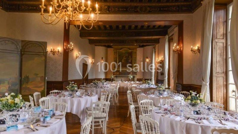 Salle de réception élégante avec tables rondes dressées, chaises blanches et lustres suspendus au plafond.