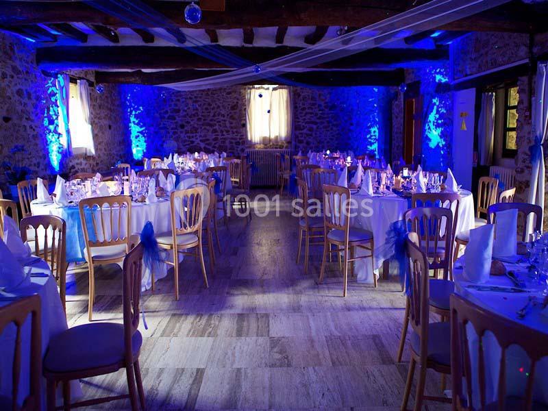 Salle de réception en pierre avec tables rondes dressées et éclairage bleu tamisé.