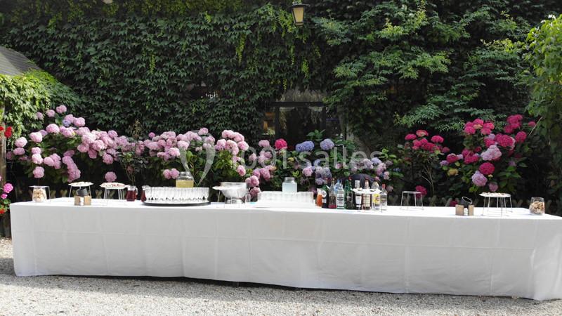 Table dressée en extérieur avec nappes blanches, boissons et verres, entourée de buissons fleuris d'hortensias.