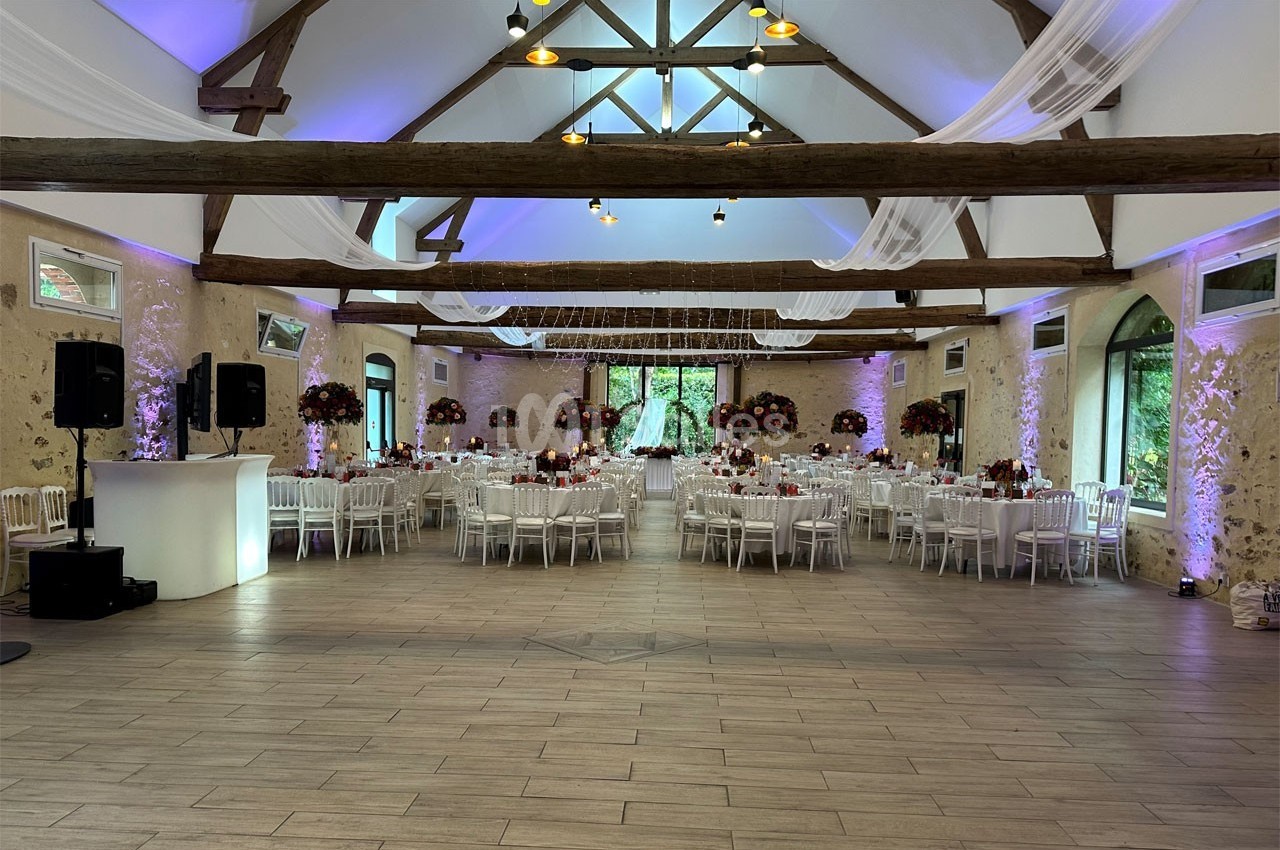 Salle de réception décorée pour un événement, avec tables rondes, chaises blanches et poutres apparentes.