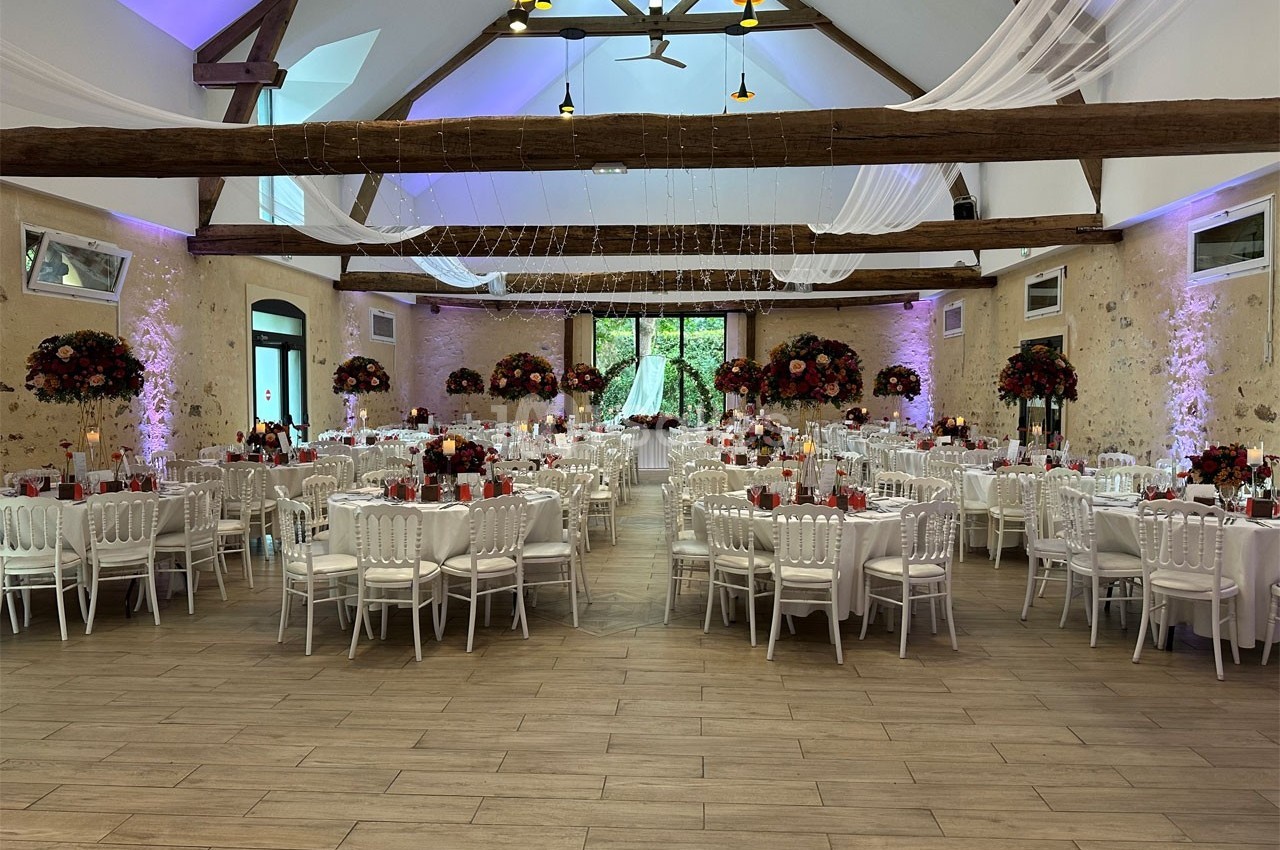 Salle de réception décorée avec des tables rondes, chaises blanches, fleurs rouges et éclairage tamisé.