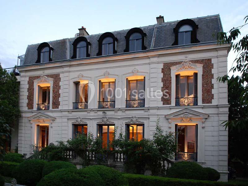Façade d'une maison bourgeoise à deux étages, avec fenêtres éclairées, entourée de verdure en début de soirée.