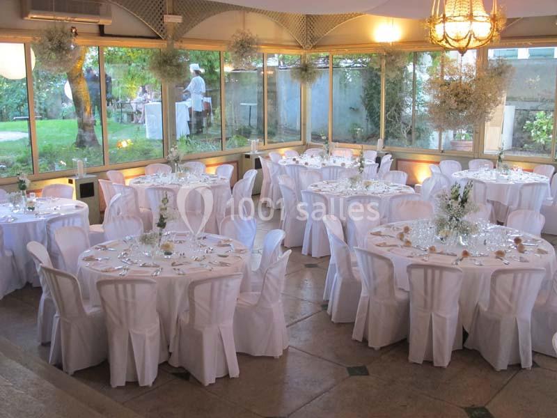 Salle de réception lumineuse avec tables rondes dressées, nappes blanches et grandes baies vitrées donnant sur un jardin.