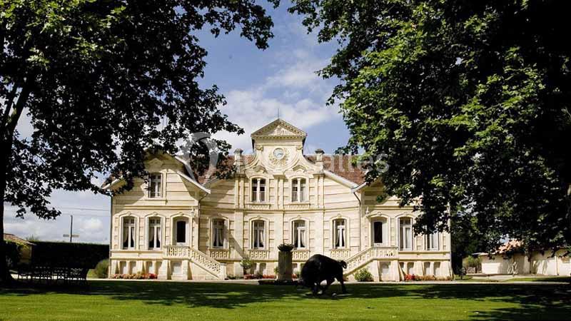 Façade d'un manoir en pierre claire entouré d'un jardin verdoyant, sous un ciel partiellement nuageux.