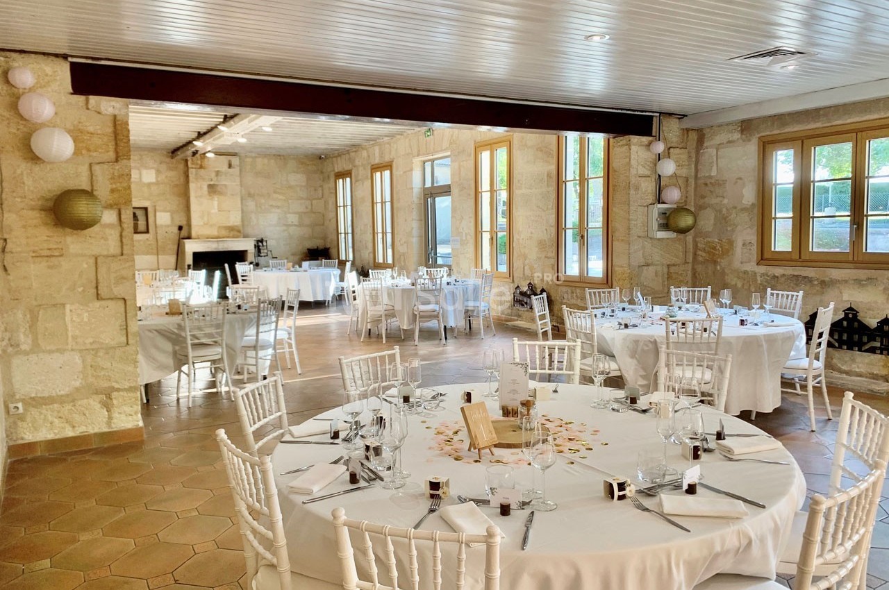 Salle de réception lumineuse avec murs en pierre, tables rondes dressées et chaises blanches, décorée pour un événement.