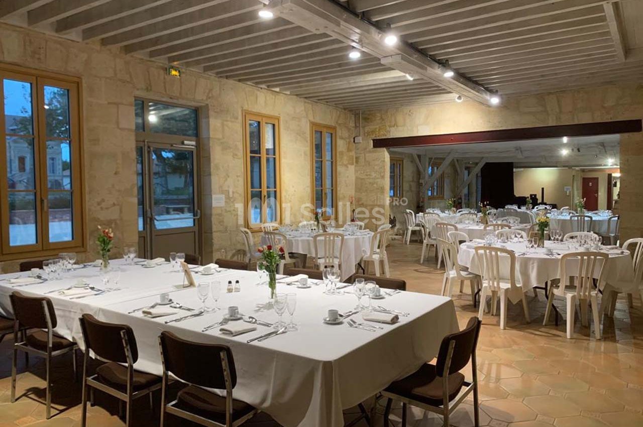 Salle de réception lumineuse avec tables dressées, nappes blanches et chaises, prête pour un événement.