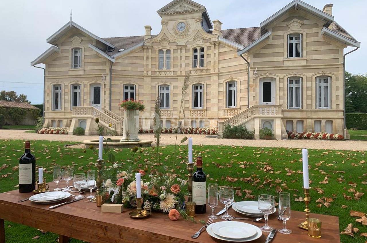 Table dressée avec vaisselle et bouteilles de vin devant un manoir en pierre entouré de pelouse et de feuilles d'automne.
