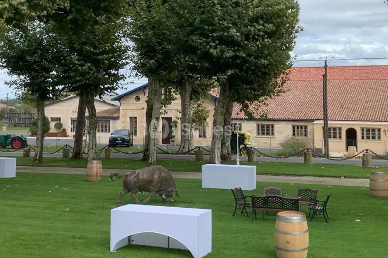 Un jardin avec des tables, des tonneaux, une statue de sanglier et des bâtiments en arrière-plan.