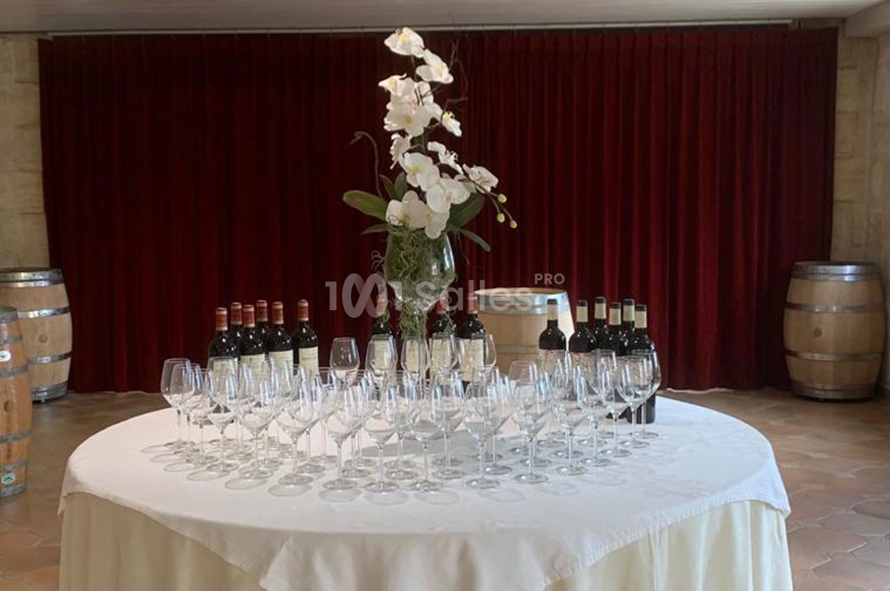 Table ronde avec nappes blanches, verres à vin alignés, bouteilles de vin et vase d'orchidées, devant un rideau rouge.