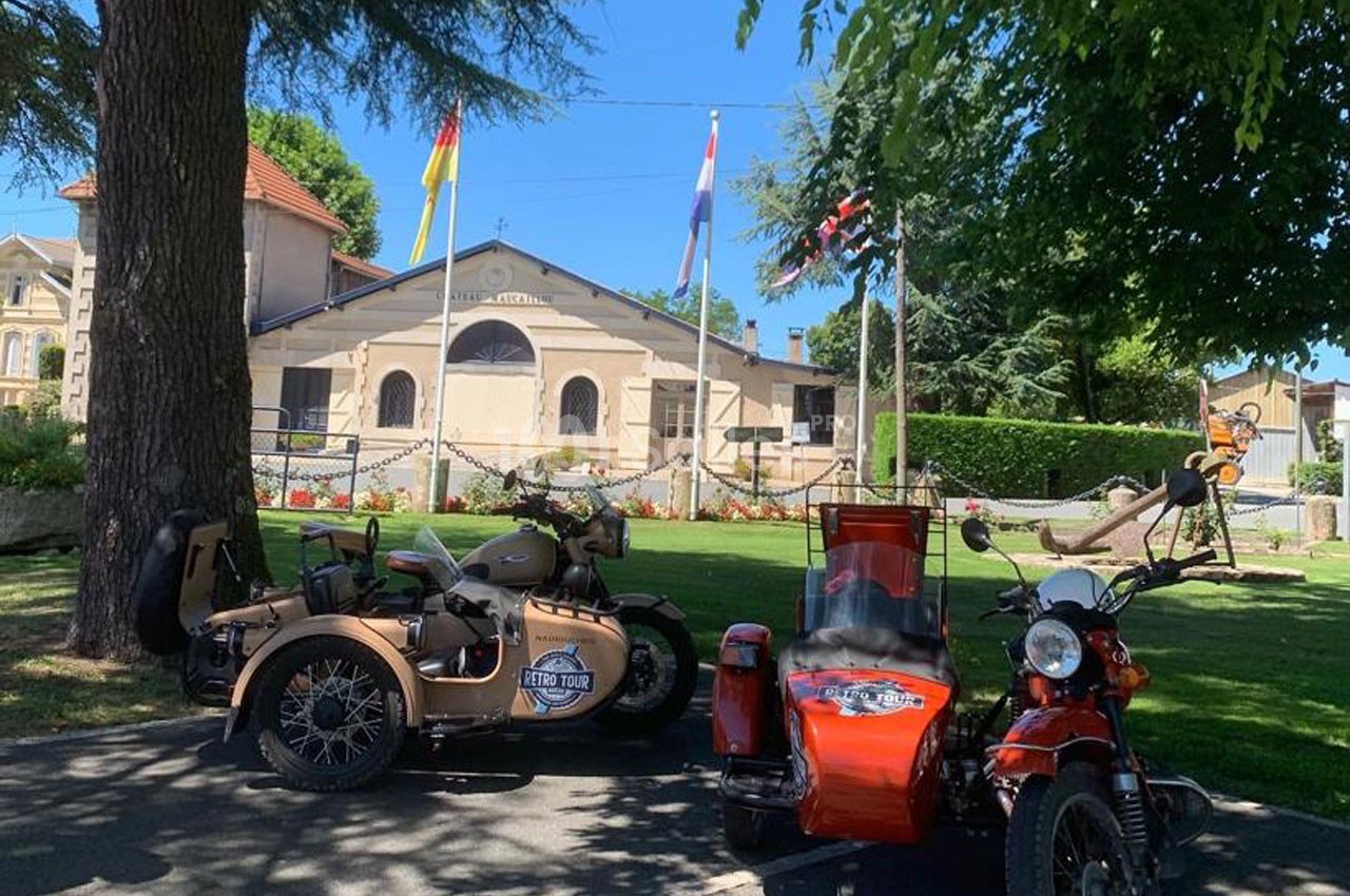 Deux motos avec side-cars stationnées devant un bâtiment ancien entouré d'arbres et de drapeaux.