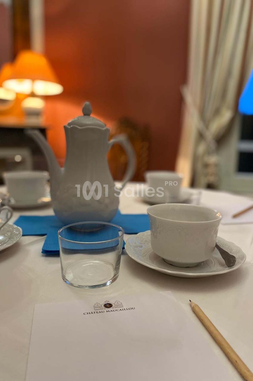 Table dressée avec une théière blanche, des tasses, un verre, un crayon et une feuille marquée ’Château Malcailou’.
