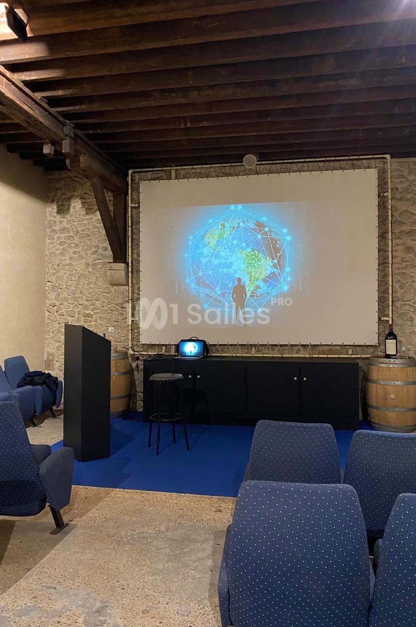 Salle de présentation avec écran projetant une image de réseau mondial, chaises bleues et tonneaux en arrière-plan.