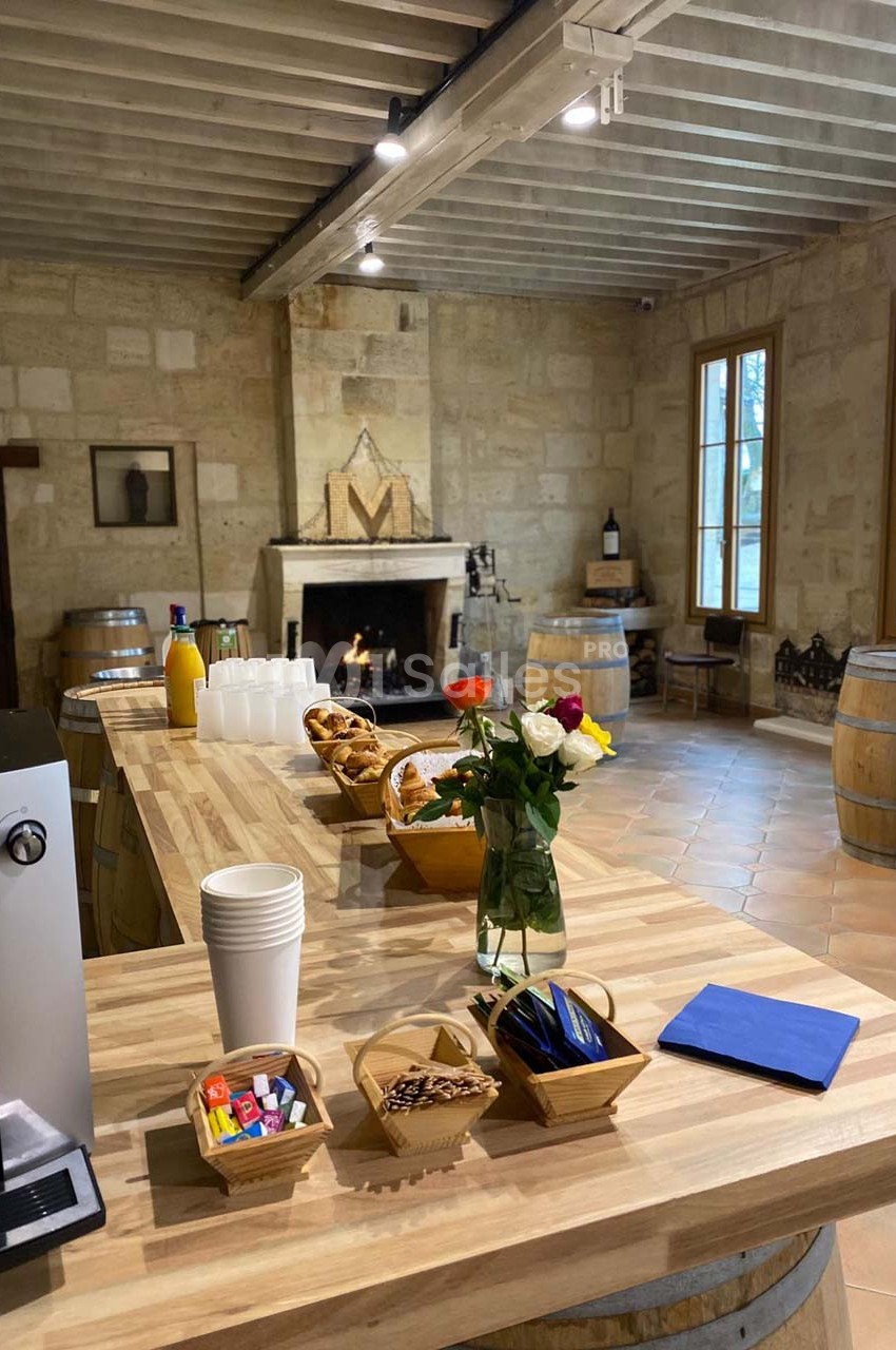 Salle rustique avec cheminée, tables en bois, barriques, et buffet comprenant viennoiseries, boissons et fleurs.