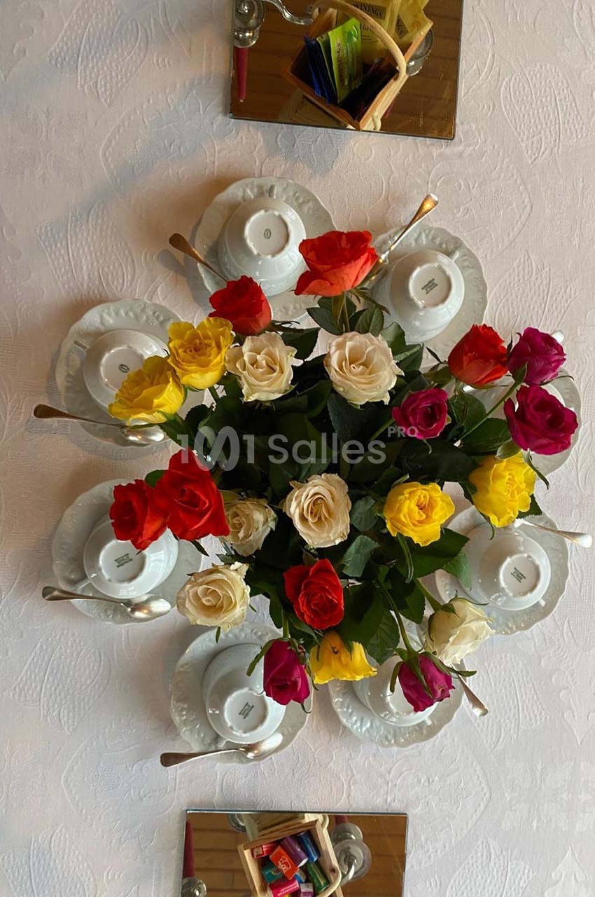 Bouquet de roses multicolores entouré de tasses blanches avec cuillères, disposé sur une nappe blanche.